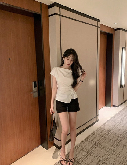Woman wearing a white cowl neck tie-waist top and black shorts in a hallway.