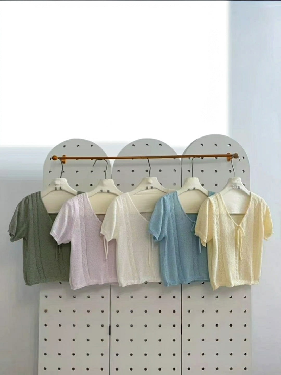 Five short-sleeved V-neck knit tops with tie-front detail in green, pink, white, blue, and yellow hanging on a pegboard