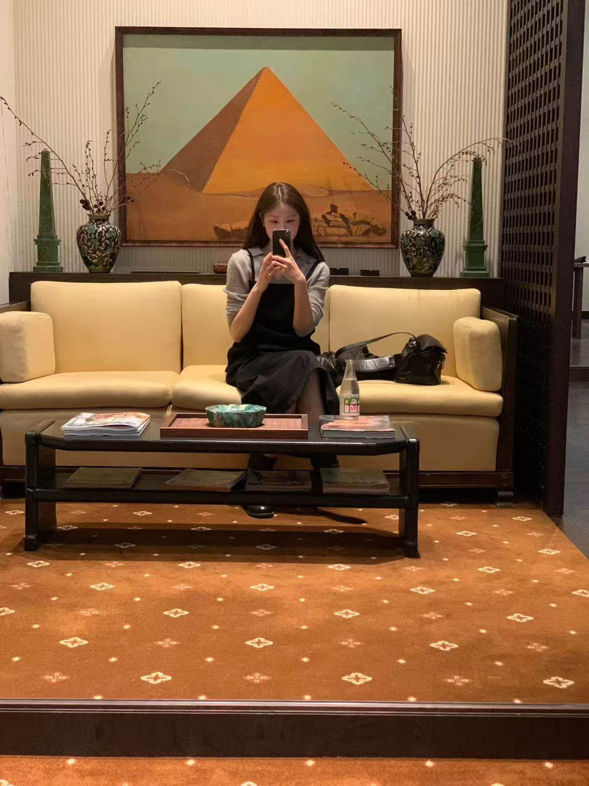 Woman taking a mirror selfie sitting on a sofa wearing a black overall dress