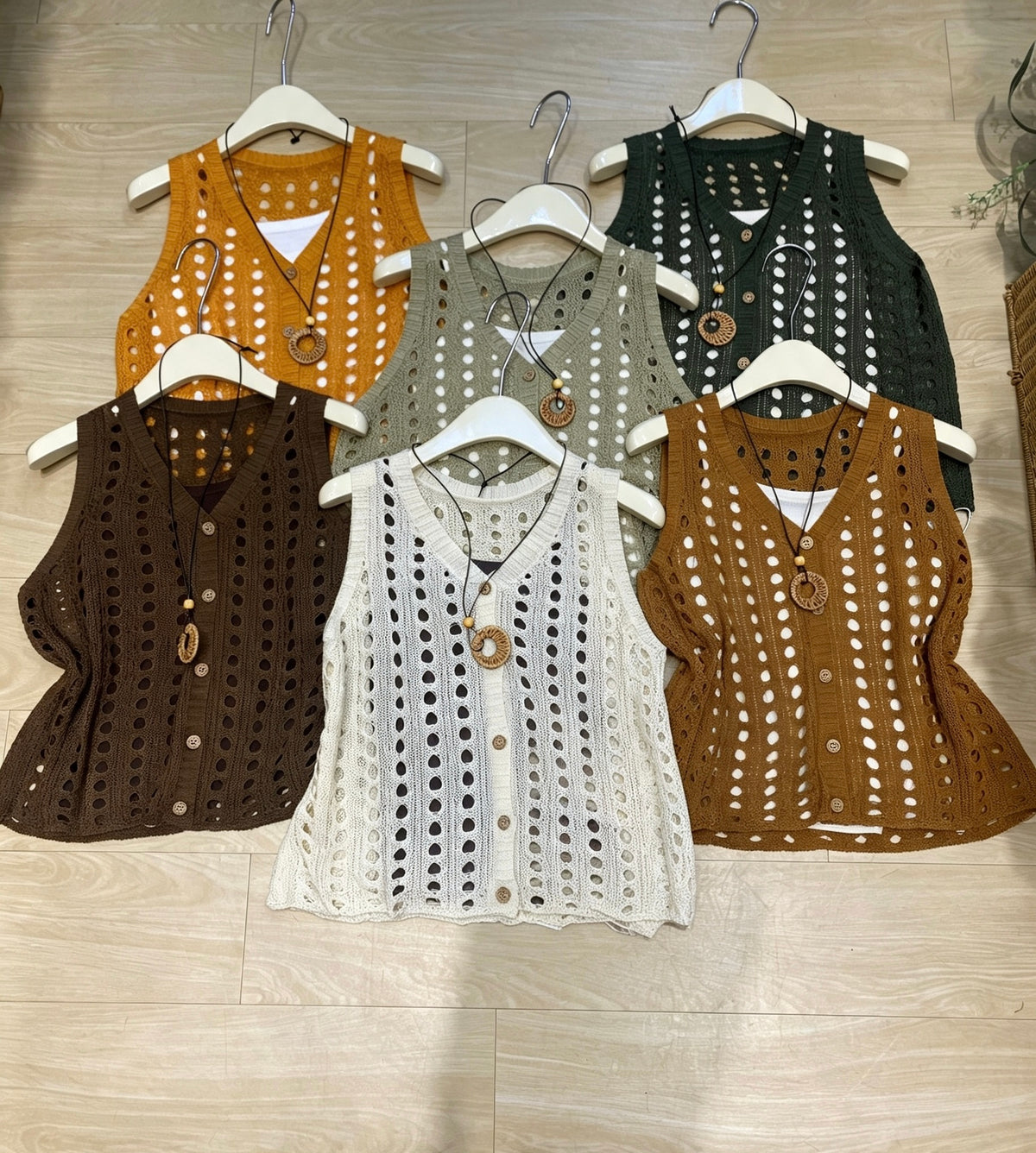 Six knitted hollow-out vests in various colors arranged on hangers on a wooden floor