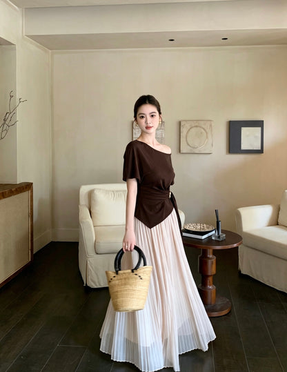 Model standing in an apricot pleated maxi skirt and brown asymmetrical top