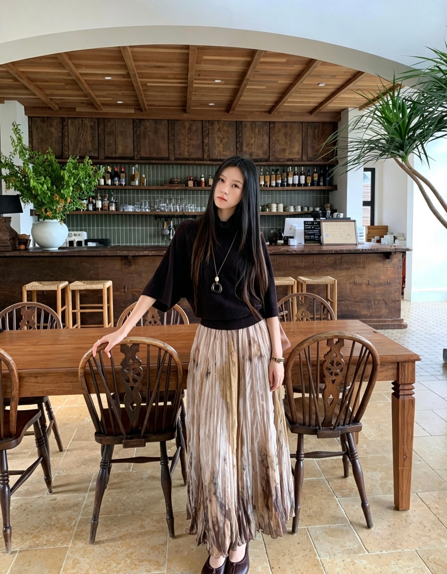 Woman in a black top and khaki tie-dye pleated skirt standing by a table