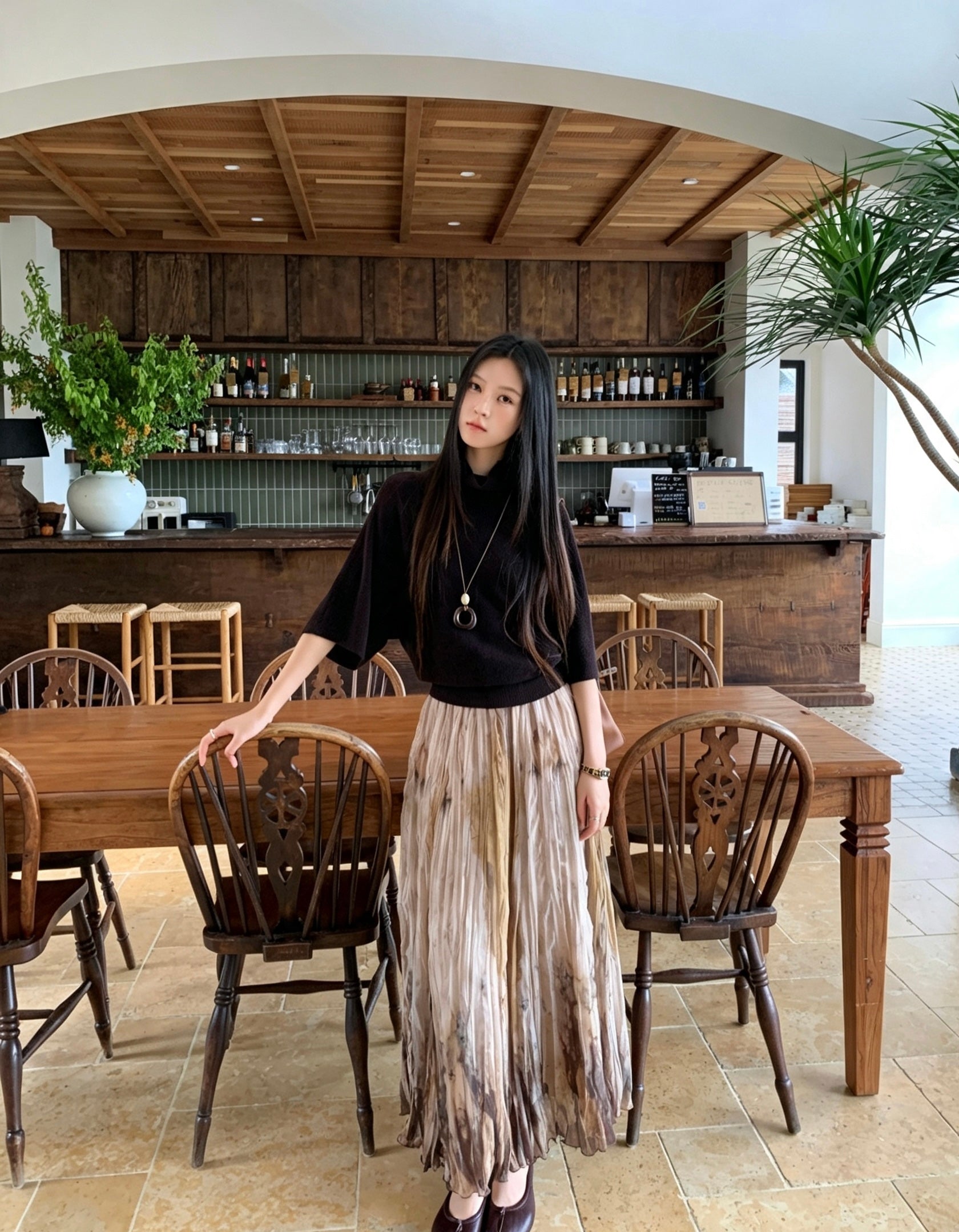 Woman in a black top and khaki tie-dye pleated skirt standing by a table