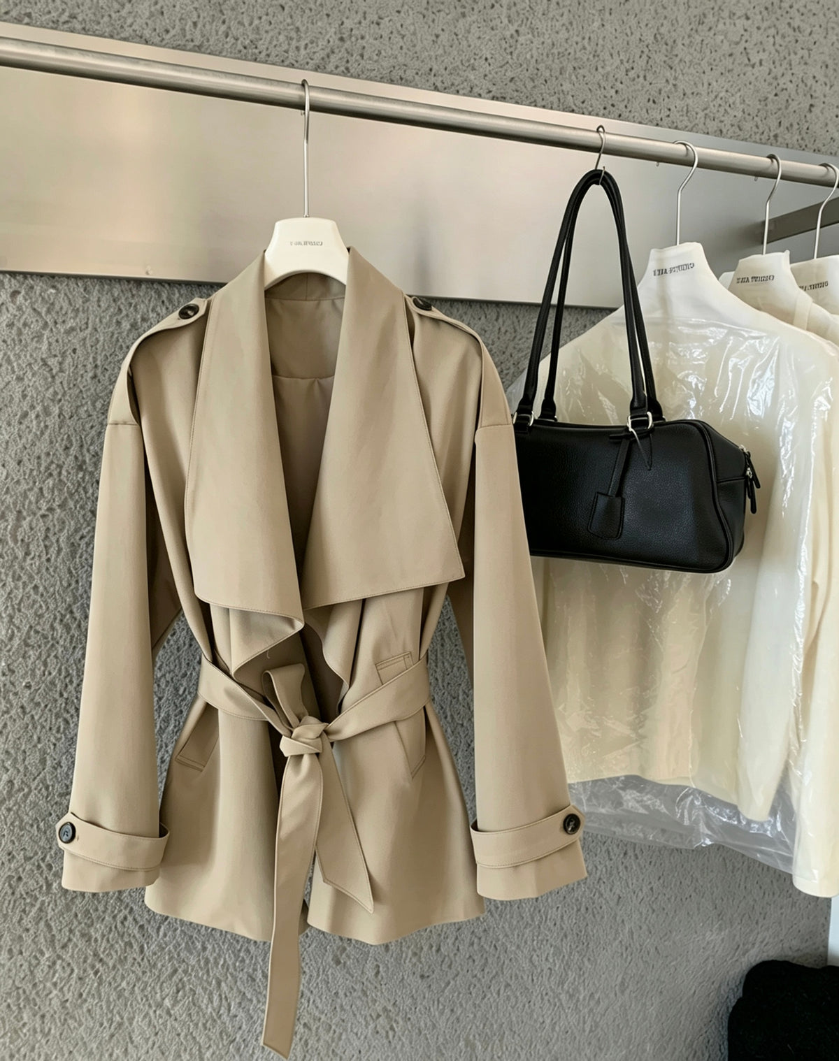 Beige trench coat with tied waist hanging on a rack next to a black bag